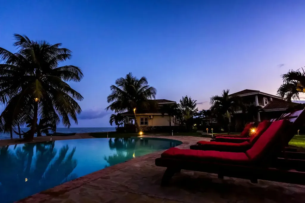 Une piscine illuminée au crépuscule, avec des transats rouges et des palmiers.