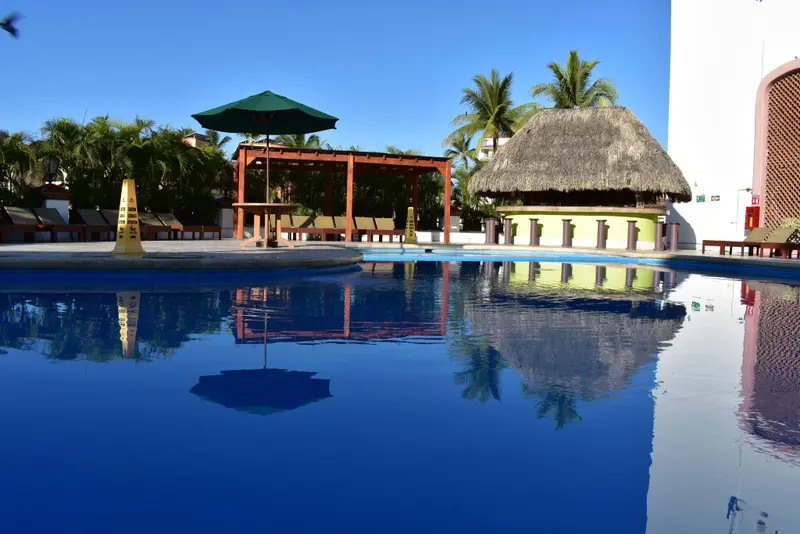 Une piscine bleu clair reflète un parasol et des palmiers sous un ciel bleu.