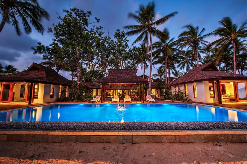 Piscine illuminée au crépuscule, entourée de bâtiments aux toits de chaume et de végétation tropicale.