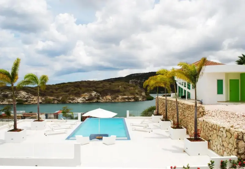 Une piscine turquoise bordée de chaises longues offre une vue sur une baie abritée et une végétation luxuriante.
