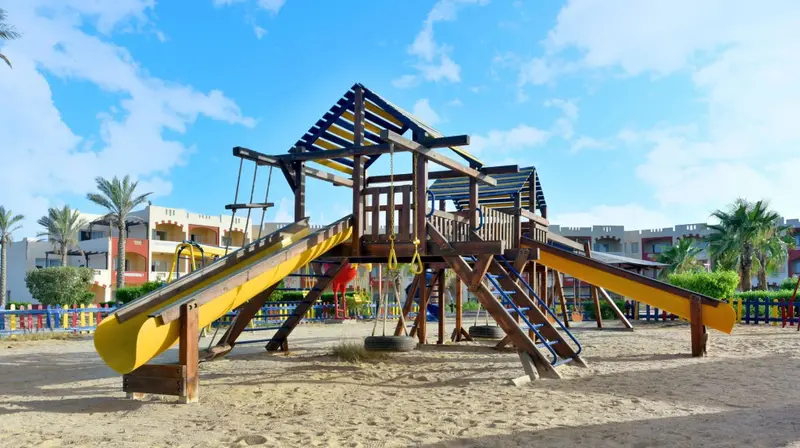 Structure de jeux pour enfants sur sol sablonneux, avec bâtiments en arrière-plan et ciel bleu.