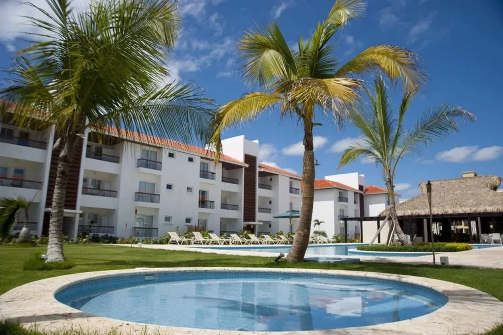 Bâtiment blanc à plusieurs étages entouré de végétation tropicale et de piscines.