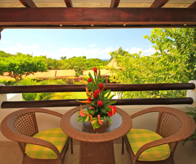 Terrasse avec table et chaises en osier, vue sur la végétation luxuriante et la mer au loin.