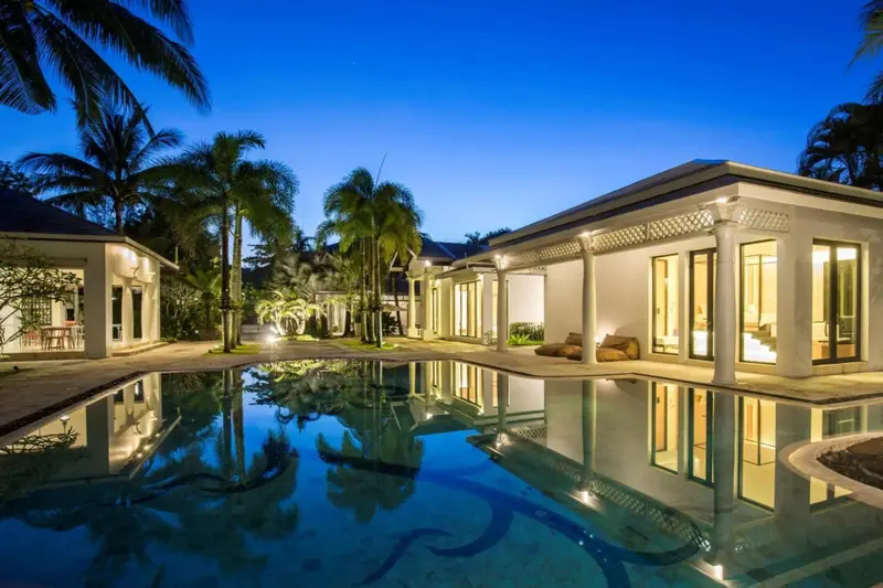 Piscine illuminée au crépuscule devant un bâtiment blanc et des palmiers.