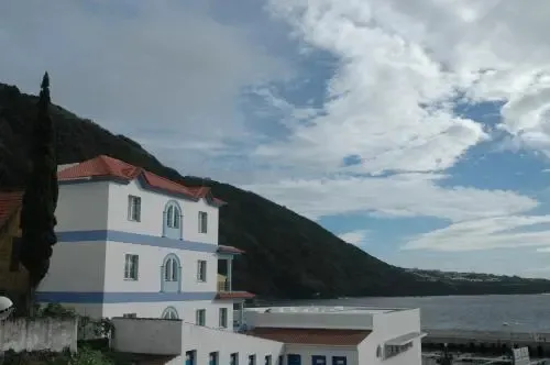 Bâtiment blanc à deux étages avec toit rouge, surplombant la mer et une colline verdoyante sous un ciel nuageux.