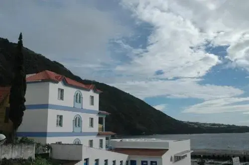 Bâtiment blanc à deux étages avec toit rouge, surplombant la mer et une colline verdoyante sous un ciel nuageux.
