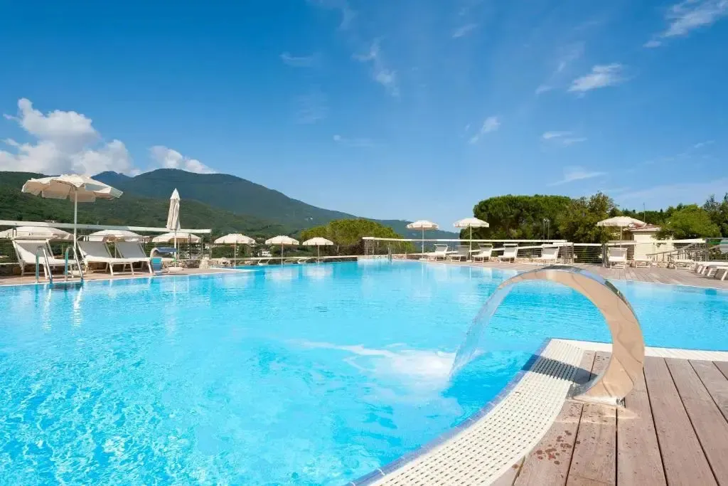 Une piscine turquoise avec des transats et des parasols, entourée de végétation et de montagnes.