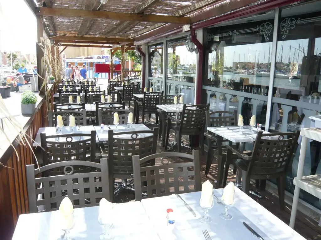 Tables et chaises disposées sur une terrasse ombragée avec vue sur un port de plaisance.