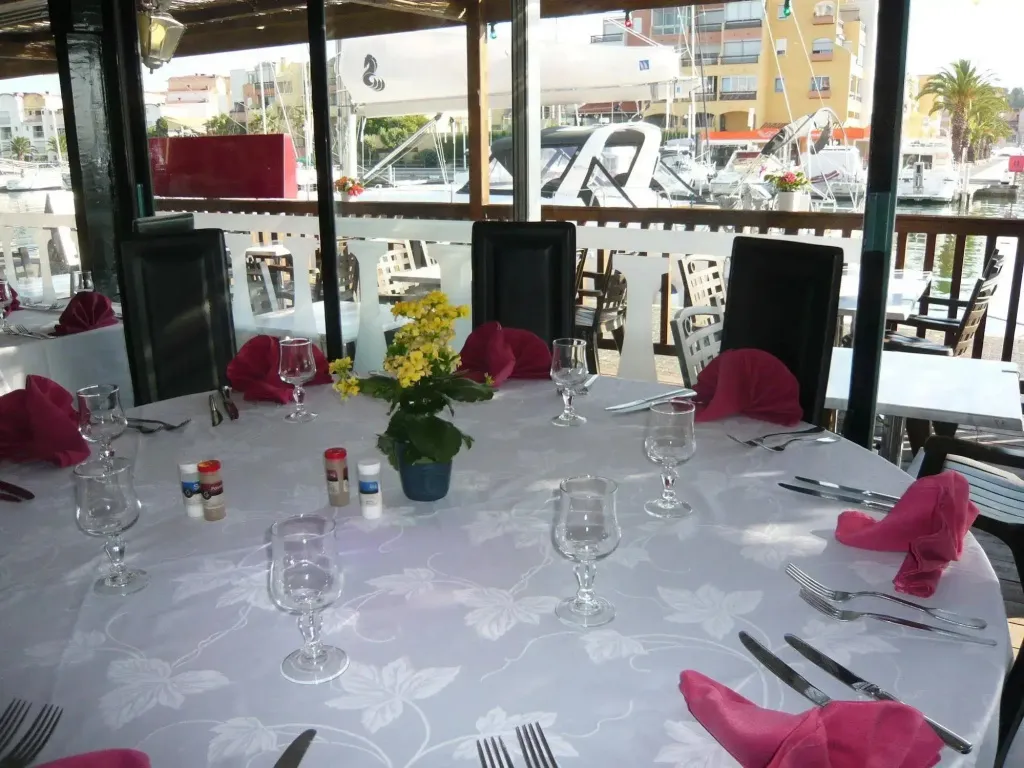 Une table dressée avec des couverts et des verres dans une salle lumineuse donnant sur un port de plaisance.