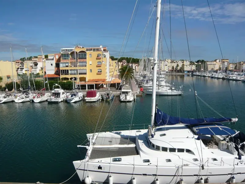 Vue d'un port de plaisance ensoleillé avec des bateaux amarrés et des bâtiments colorés en arrière-plan.