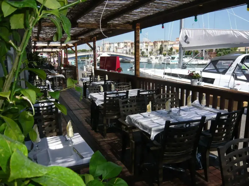 Terrasse ombragée avec tables nappées et vue sur un port de plaisance.