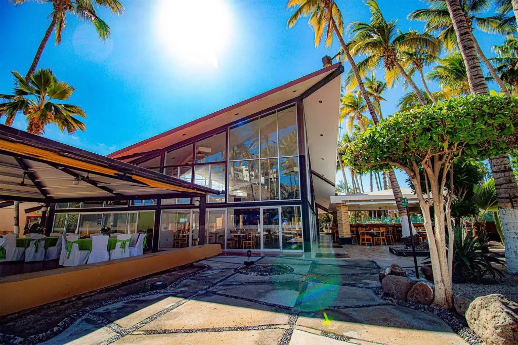 Bâtiment moderne aux grandes baies vitrées entouré de végétation tropicale et d'une terrasse ombragée.
