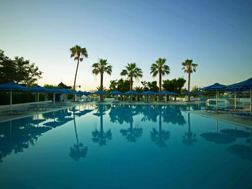 Une piscine rectangulaire bordée de transats et d'ombrelles bleues, avec des palmiers en arrière-plan.