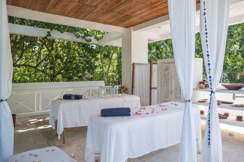 Deux tables de massage blanches sont disposées dans un espace extérieur ombragé et décoré de fleurs.