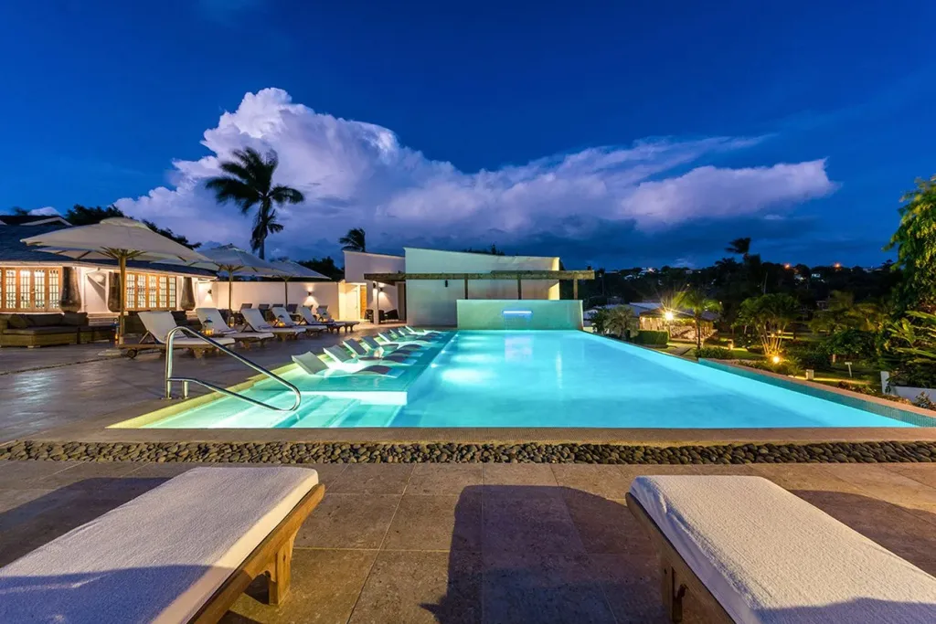 Piscine illuminée avec transats et végétation luxuriante sous un ciel nocturne.