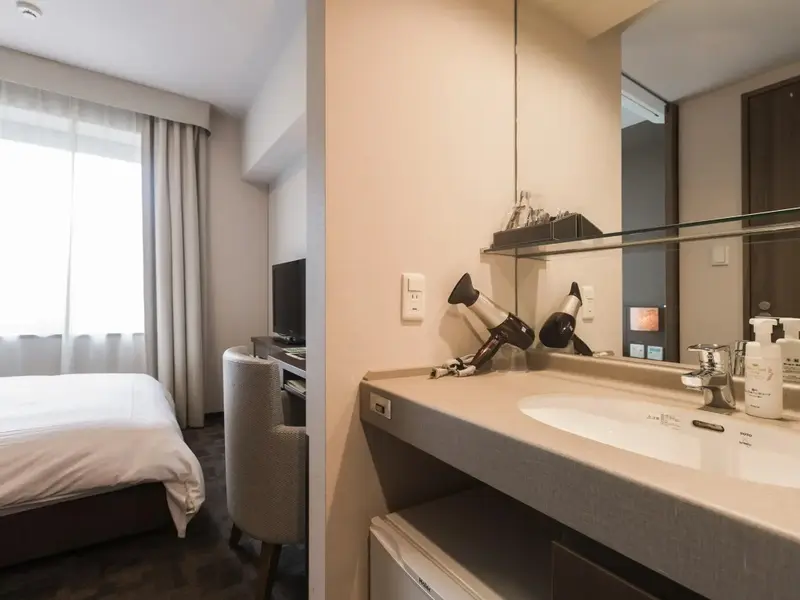 Une salle de bain lumineuse avec un lavabo et un sèche-cheveux, donnant sur une chambre.