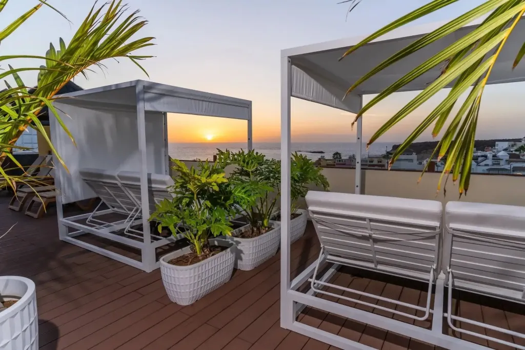 Terrasse aménagée avec des transats blancs et des plantes vertes, face à la mer au coucher du soleil.