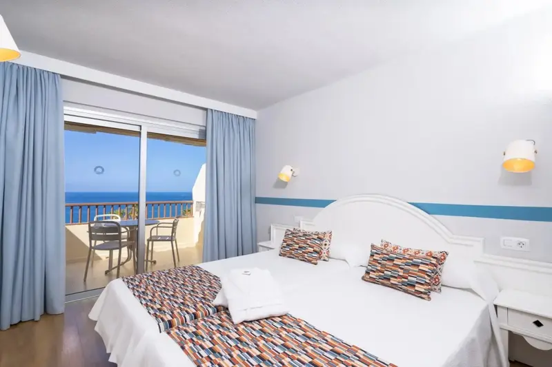 Chambre lumineuse avec un grand lit et une vue sur la mer depuis un balcon.