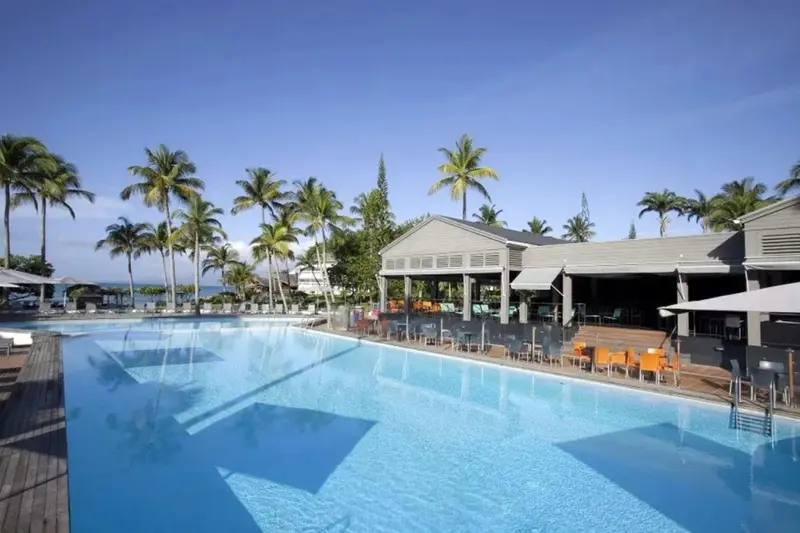 Une grande piscine turquoise bordée de chaises longues et de palmiers sous un ciel bleu.