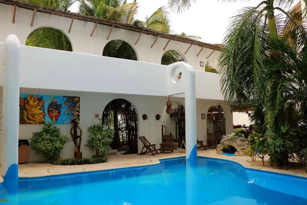 Une piscine turquoise au premier plan, bordée d'une terrasse blanche et de végétation tropicale.