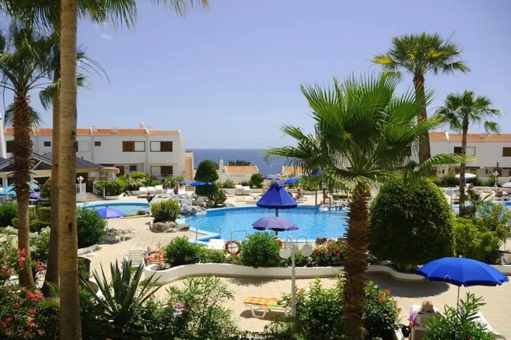 Une piscine ensoleillée avec des transats et des parasols, entourée de végétation luxuriante et de bâtiments blancs.