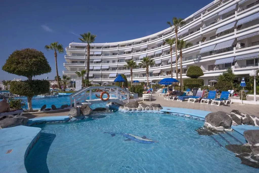 Piscine turquoise avec pont et transats bleus devant un bâtiment blanc.