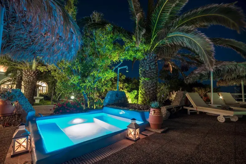 Une piscine éclairée au milieu d'une végétation luxuriante, avec des transats à proximité.