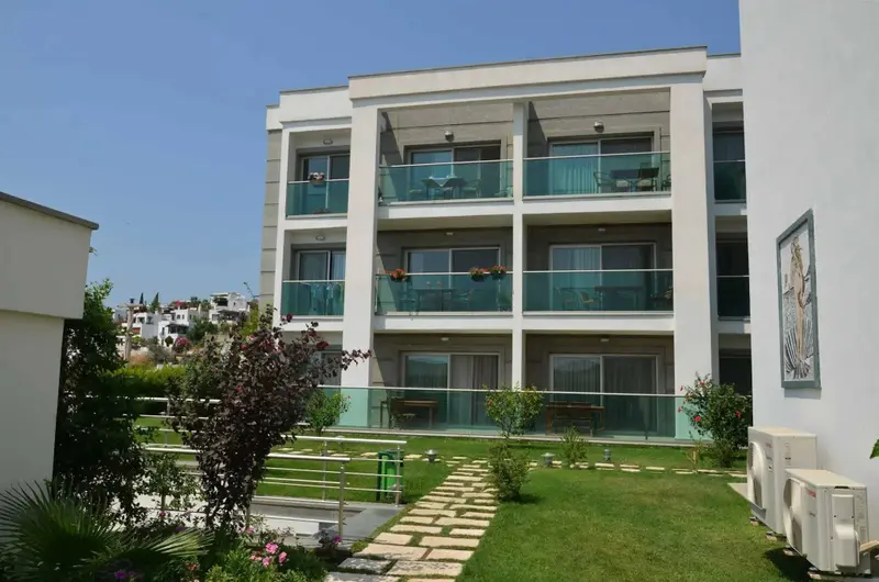 Bâtiment blanc à plusieurs étages avec des balcons fleuris et un jardin soigné.