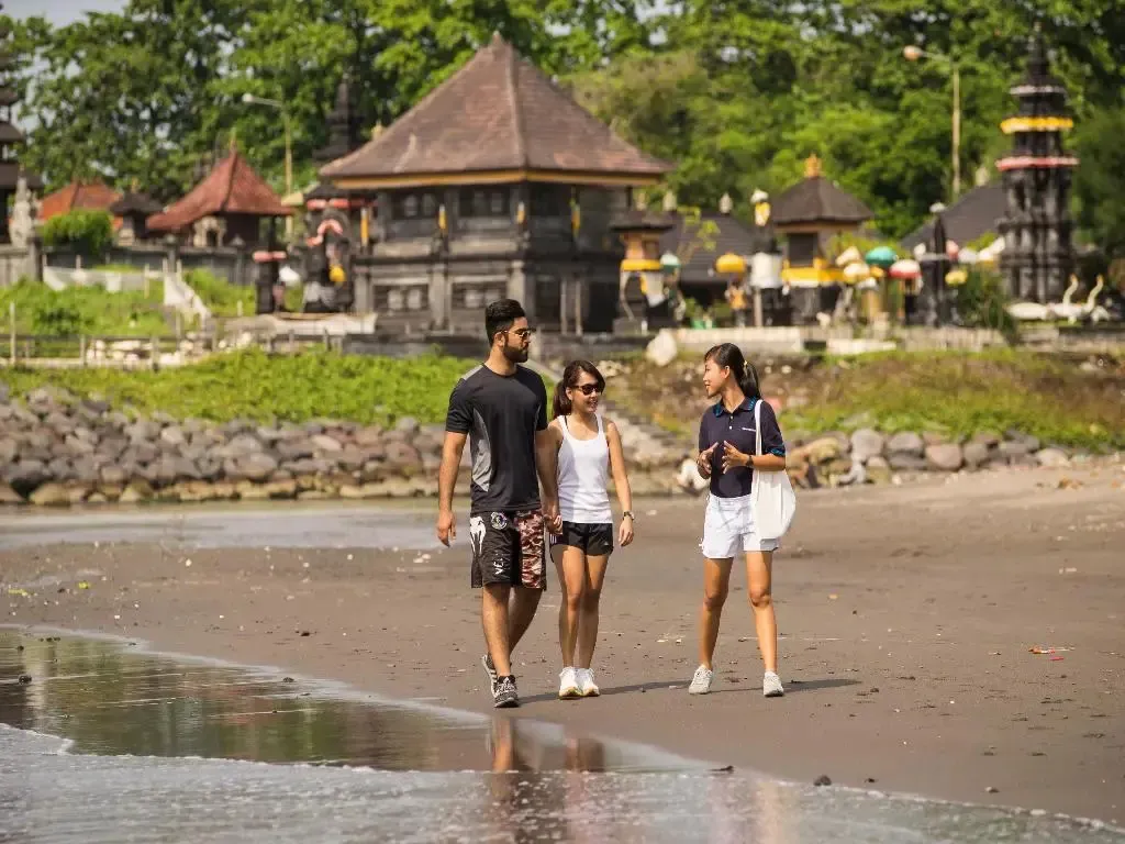 Trois personnes se promènent sur une plage de sable gris face à des temples traditionnels.