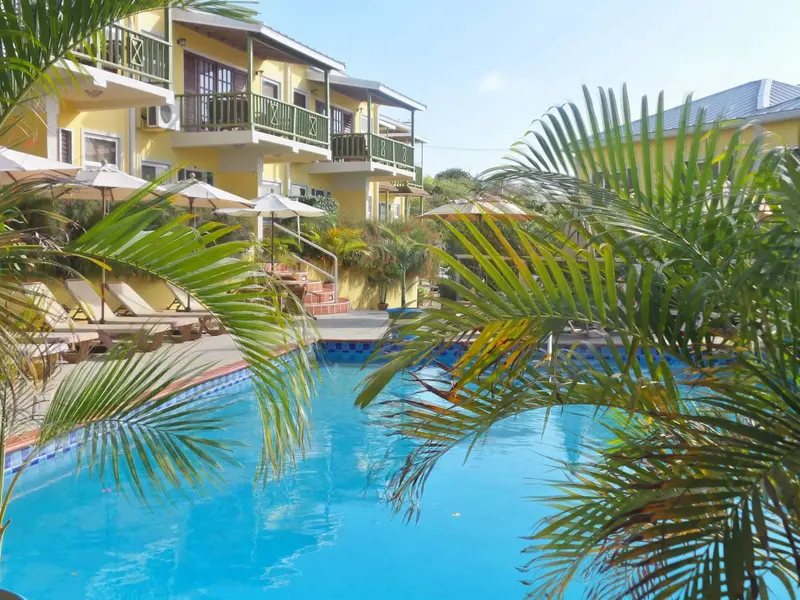 Une piscine turquoise bordée de transats et de parasols, devant un bâtiment jaune à balcons.