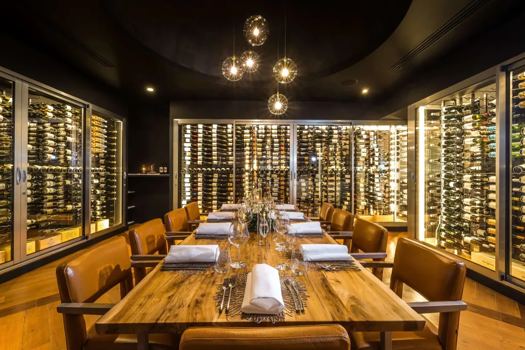 Une longue table en bois est mise en valeur par des luminaires suspendus dans une salle entourée de caves à vin.