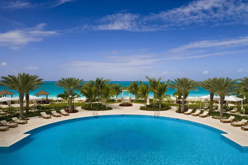 Piscine turquoise bordée de transats, avec une plage de sable fin et des palmiers en arrière-plan.