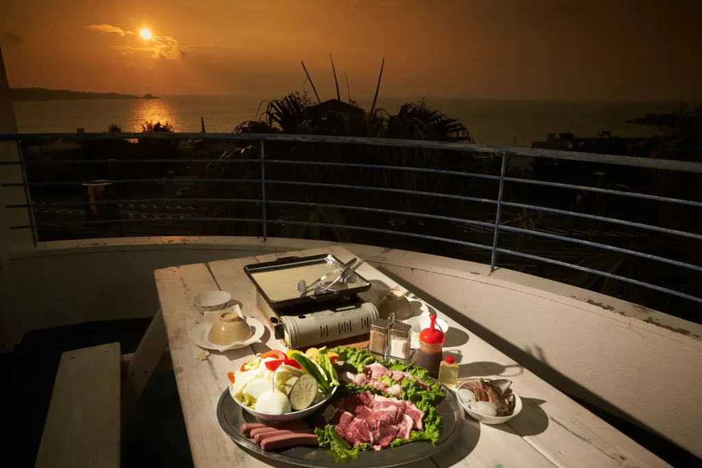 Une table garnie de mets variés est mise en scène sur une terrasse avec vue sur la mer au coucher du soleil.