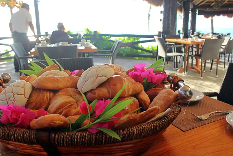 Une corbeille de viennoiseries est présentée sur une table en bois, avec une vue sur une terrasse et la mer en arrière-plan.