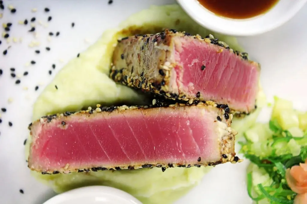 Assiette de poisson rouge en tranches, recouvert de graines noires, servi sur une purée verte et accompagné d'une salade.
