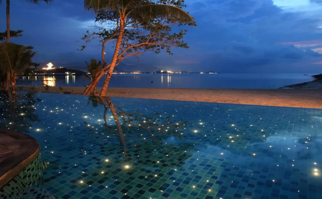 Une piscine à débordement reflète les lumières du rivage au crépuscule.