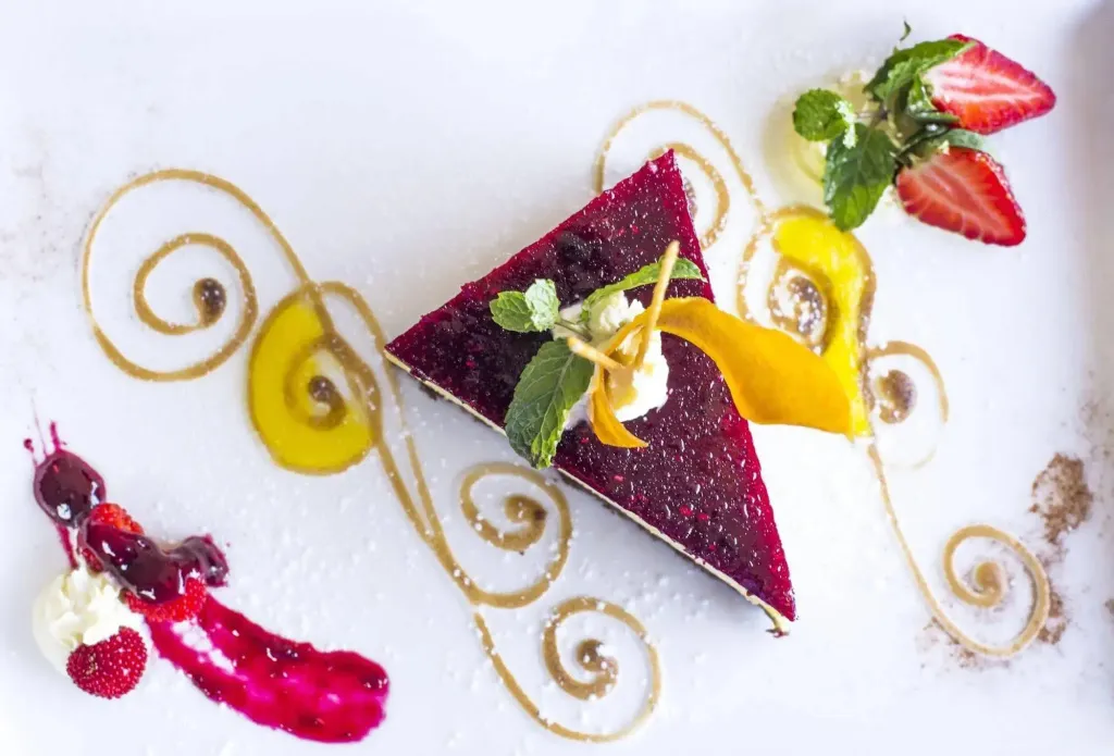 Une part de gâteau rouge décorée de fruits frais et de motifs dorés est présentée sur une assiette blanche.