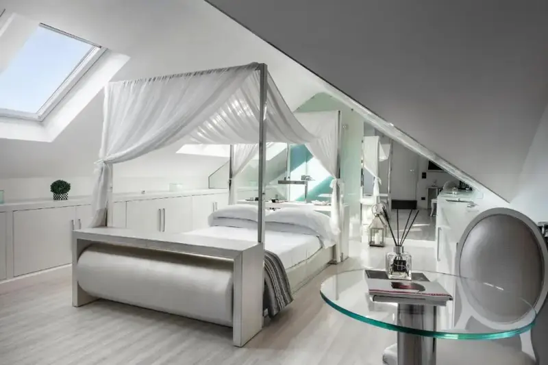 Une chambre mansardée lumineuse avec un lit à baldaquin et une table basse en verre.