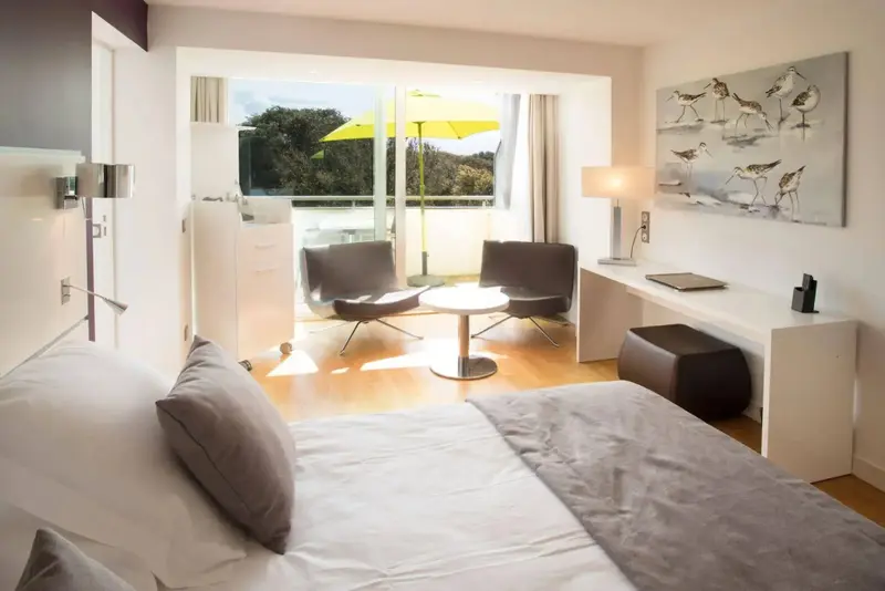 Une chambre lumineuse s'ouvre sur un espace extérieur avec des fauteuils et un parasol.