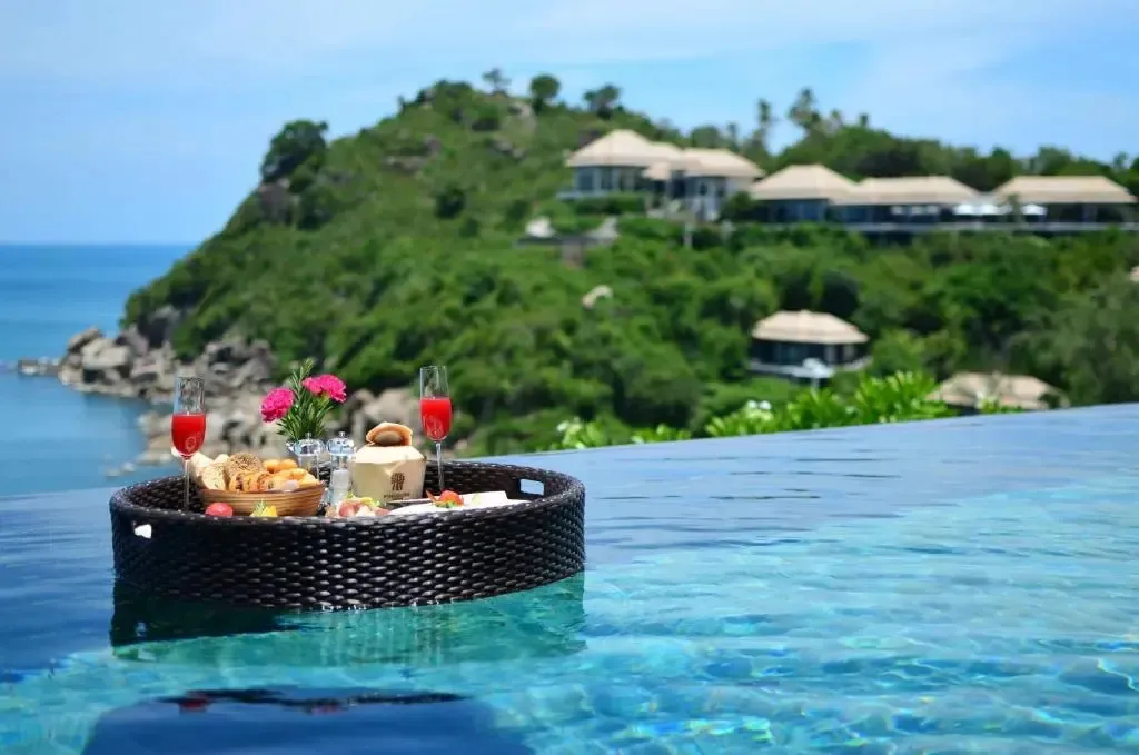 Un plateau de nourriture et de boissons flotte sur une piscine à débordement avec vue sur la mer et la végétation.