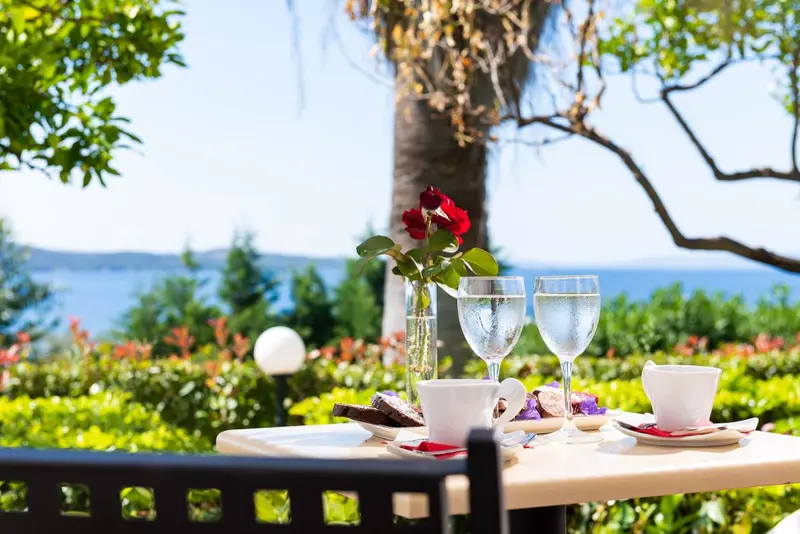 Une table est dressée avec des tasses, des pâtisseries et des verres d'eau devant un paysage verdoyant et une vue sur la mer.