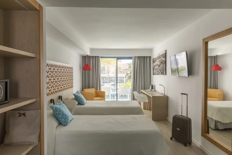 Une chambre lumineuse avec deux lits simples et un bureau près d'une baie vitrée.
