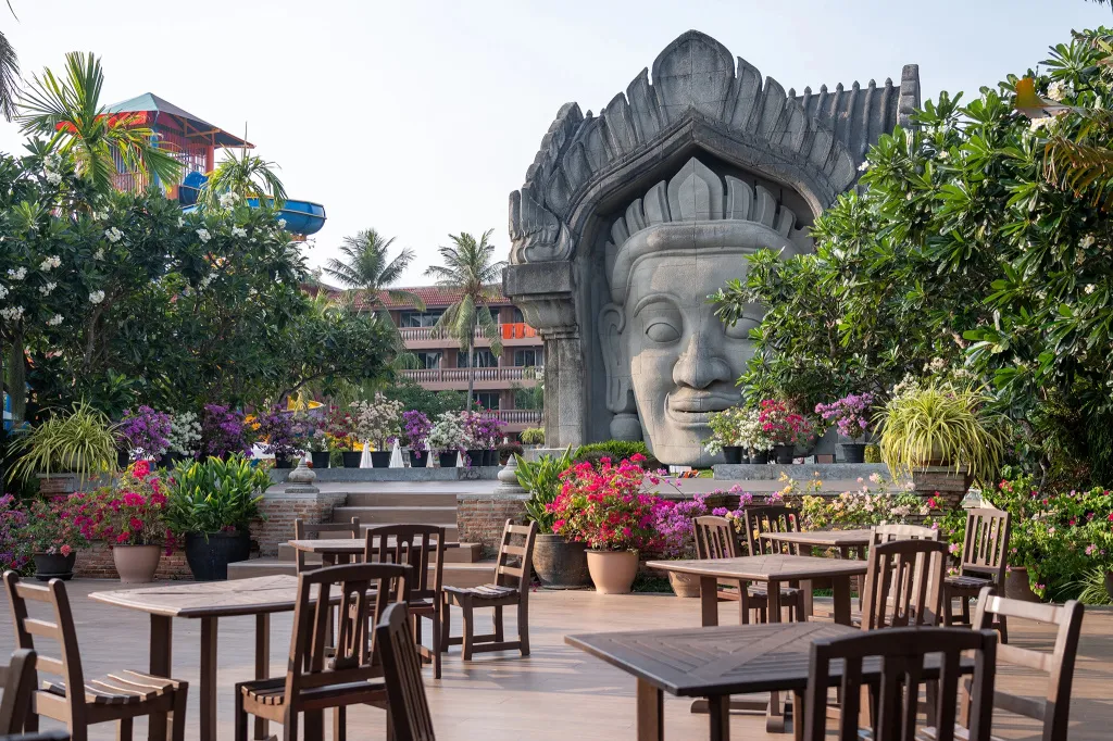 Tables et chaises en bois disposées sur une terrasse ombragée, devant une grande sculpture de visage.