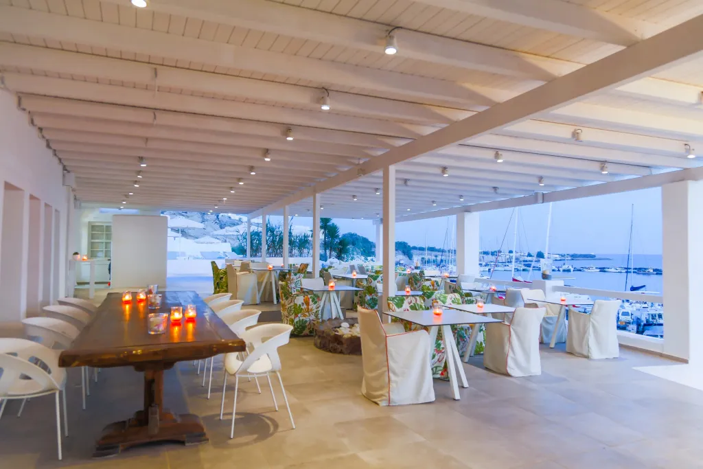 Une longue table en bois est dressée dans un espace extérieur couvert avec vue sur la mer et un port de plaisance.