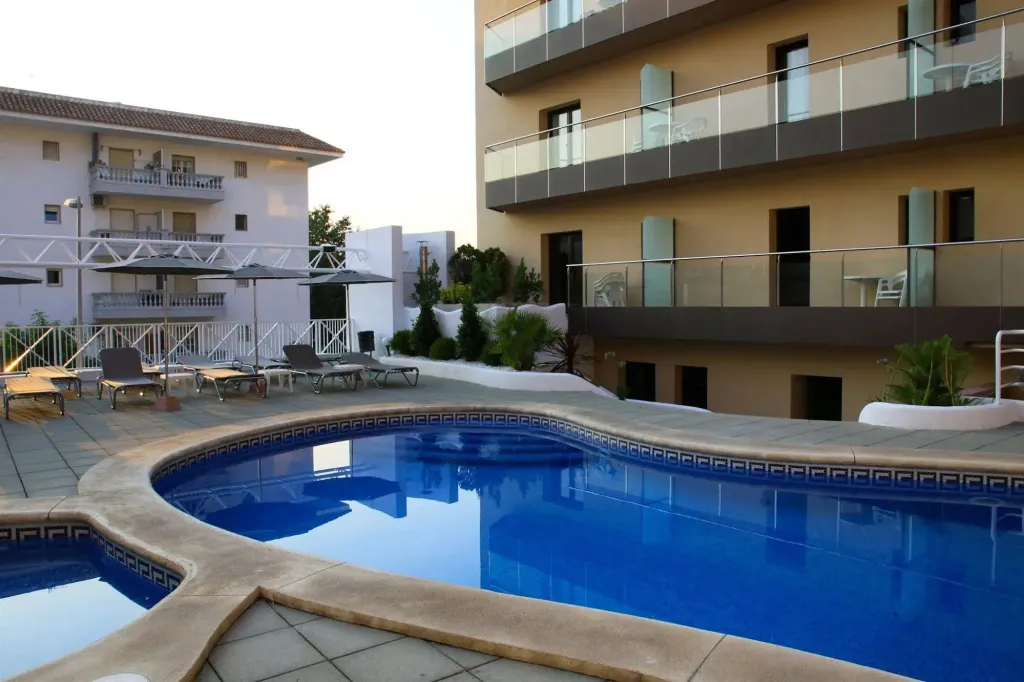 Une piscine rectangulaire bordée d'une terrasse avec des transats et des parasols, devant un bâtiment à plusieurs étages avec des balcons.