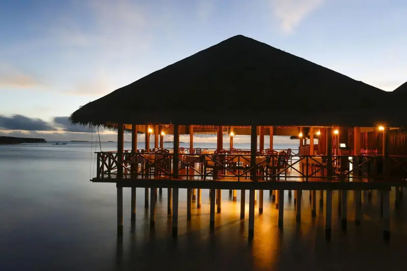 Un restaurant sur pilotis illuminé au crépuscule surplombe l'océan calme.