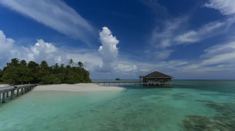 Une plage de sable blanc mène à une structure sur pilotis au-dessus de l'eau turquoise.