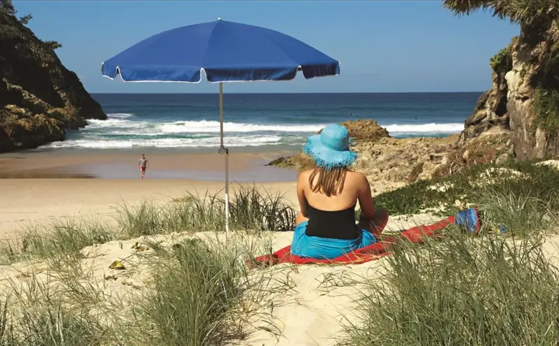 Une personne assise sur une serviette de plage sous un parasol bleu regarde la mer.