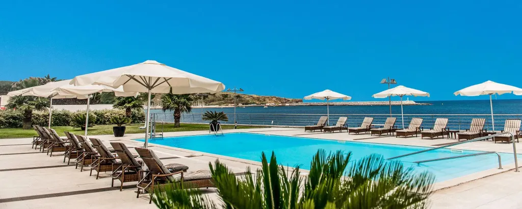 Transats blancs disposés autour d'une piscine turquoise face à la mer.
