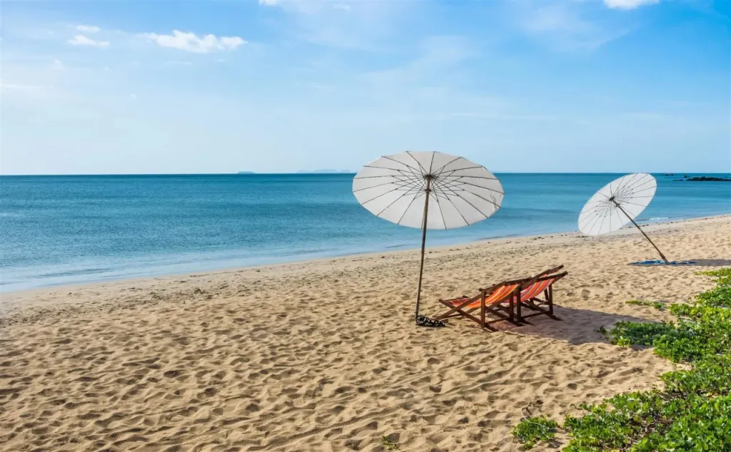 Deux transats et parasols sur une plage de sable fin bordée par une mer turquoise.
